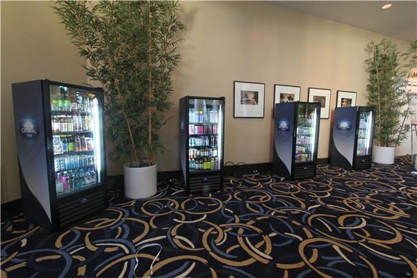 Four glass-front Crystal Cooler refrigerators stocked with bottled drinks line a wall between potted bamboo plants above a patterned carpet.