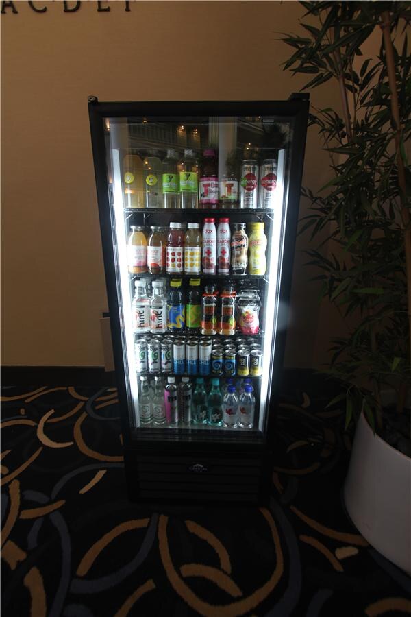 A lit glass-front refrigerator holds shelves of bottled and canned drinks, including Hint water bottles, beside a potted plant on a patterned carpet.