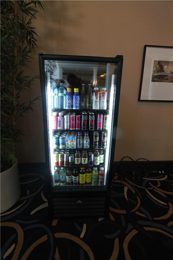 A tall glass-door refrigerator lit from inside, stocked with bottled and canned drinks including Zico bottles on the top shelf, standing on a patterned carpet beside a potted plant.
