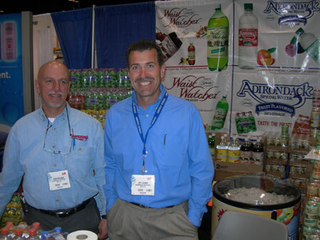Two men in blue shirts with name badges stand in a booth with Adirondack Spring Water and Waist Watchers signs and stacked bottles behind them.
