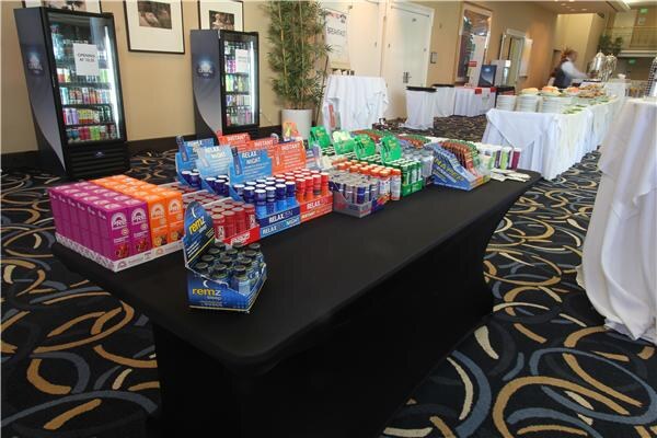 A long black-draped table covered with boxes and displays of energy and relaxation shots, including a Remz display, with glass-door coolers and catering tables in the background.