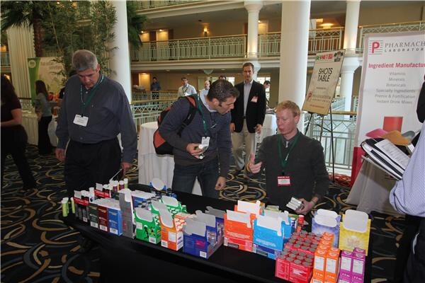 Three men with lanyards stand around a table of boxed shot products. A Shots Table sign and a Pharmachem Laboratories banner stand behind them, with a balcony in the background.