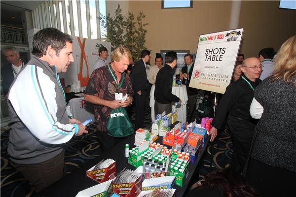 Attendees crowd around a table of shot products beside a Shots Table sign sponsored by Pharmachem Laboratories. One man holds a small bottle and another wears a green tote bag.