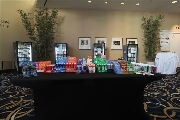 A wide view of a black-draped table lined with boxes of shot products, including a Remz display, in front of four glass-door coolers and a white-draped table in a hotel ballroom.