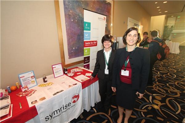 Two women with name badges stand smiling beside a table with a red cloth bearing the Symrise logo. Brochures and sample items sit on the table, and a Symrise banner stands behind.
