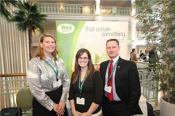Two women and a man wearing green lanyards and name badges pose together in front of a green Wild banner that reads That certain something, with a palm and balcony behind them.