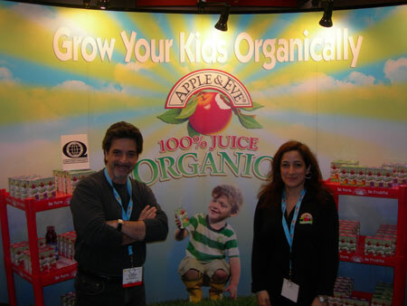 A man and a woman wearing lanyards stand in the Apple & Eve booth in front of a backdrop reading 'Grow Your Kids Organically' with a child holding a juice box, and shelves of juice boxes on both sides.