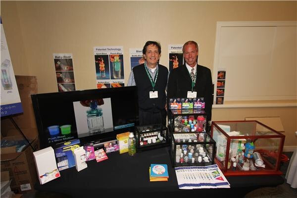 Two men with lanyards stand behind a table displaying BlastCap bottle cap products in glass cases, with packaging and brochures in front and posters on the wall behind.