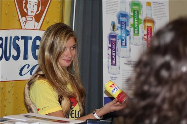 A woman with long blonde hair in a yellow shirt holds a yellow Bustelo Cool can. A Bustelo Cool sign is at left and a BlastMax poster with colorful bottles is behind her.