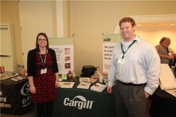 A woman in a red patterned dress and a man in a light blue shirt stand beside a green Cargill-draped table with brochures and banners; another person sits at a table behind them.