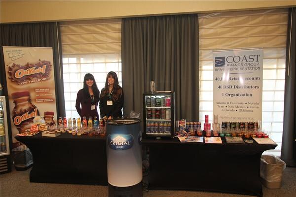 Two women with long dark hair stand behind black tables of bottled drinks, with an Oh Yeah! banner, a Coast Brands Group banner, a Crystal Cooler unit and a small display fridge.