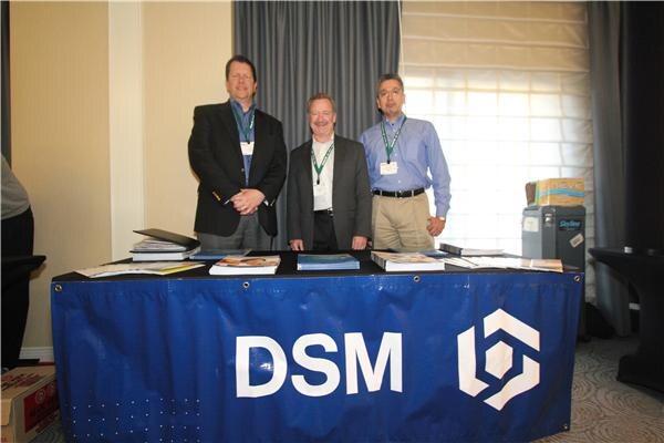 Three men in jackets and collared shirts stand behind a blue DSM-branded table holding brochures and binders, wearing green lanyards.