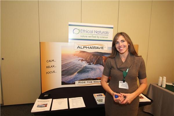 A smiling woman with long brown hair stands in front of an Ethical Naturals AlphaWave display showing a coastal cliff and the words Calm, Relax, Focus, with brochures on the table.