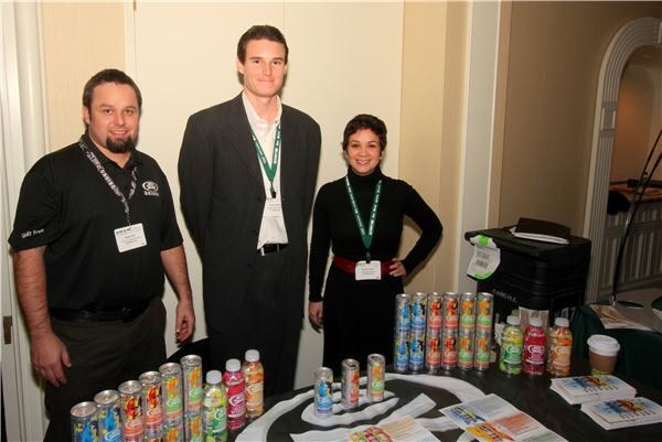 A bearded man in a black polo, a tall man in a pinstripe suit and a woman in black stand behind a table of eX canned and bottled drinks and printed materials.