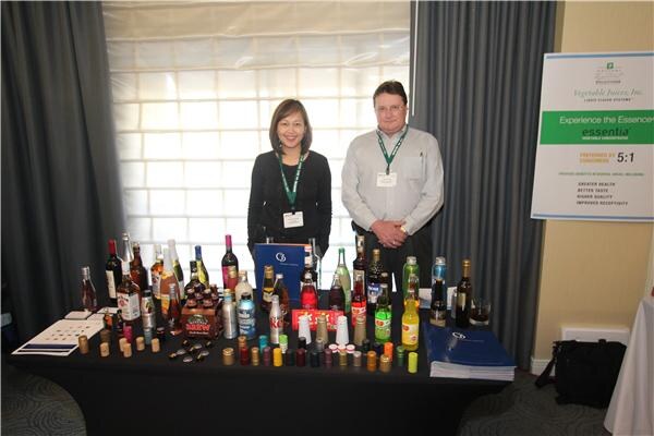 A woman and a man stand behind a black table filled with bottles and small vials; a Vegetable Juices, Inc. Essentia sign at right reads Experience the Essence.