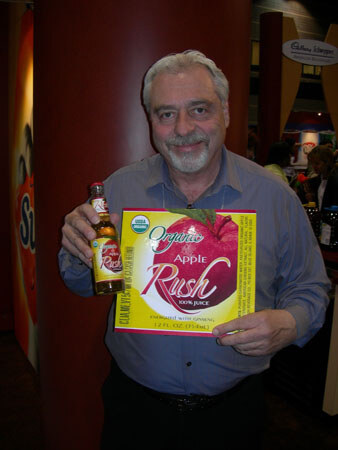 A gray-haired, bearded man in a blue shirt smiles while holding a small bottle and a box of Organic Apple Rush 100% juice enriched with ginseng.