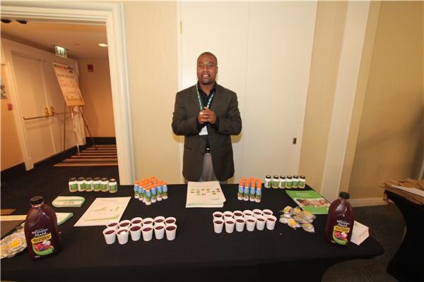 A man in a dark blazer stands behind a black table with Minute Maid juice bottles, small sample cups, and rows of small bottles and printed sheets.