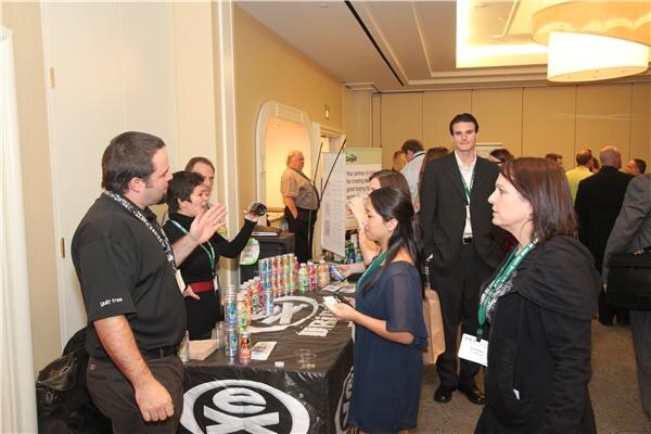 A crowd of attendees gathers around a black table of eX drinks, where a man in a black polo gestures while talking with several people wearing lanyards.