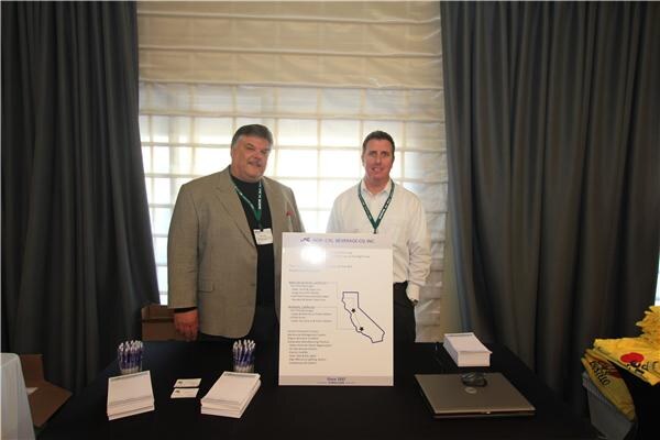Two men wearing lanyards stand behind a draped table with brochures and a Nor-Cal Beverage Co. Inc. sign showing a map of California.