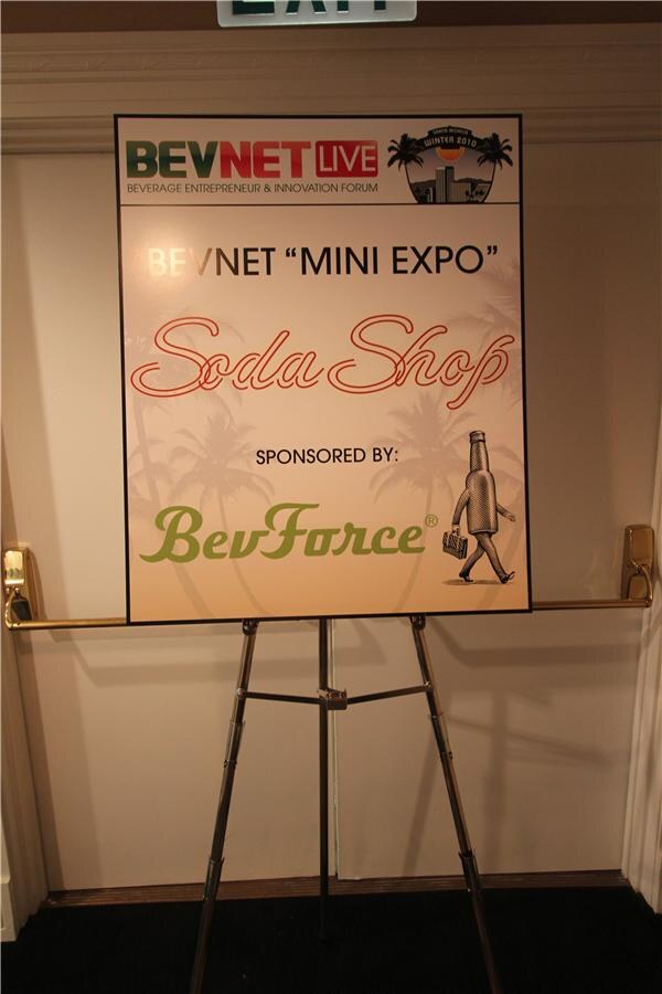 Easel sign reading BevNET Mini Expo Soda Shop, sponsored by BevForce, with a cartoon bottle carrying a briefcase and the BevNET Live Winter 2010 logo.