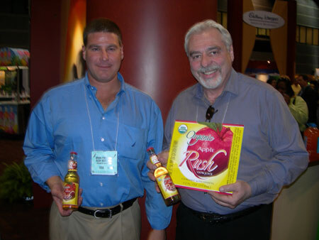 Two smiling men stand together holding Apple Rush bottles. The man on the right, gray-haired and bearded, also holds a box of Organic Apple Rush juice.
