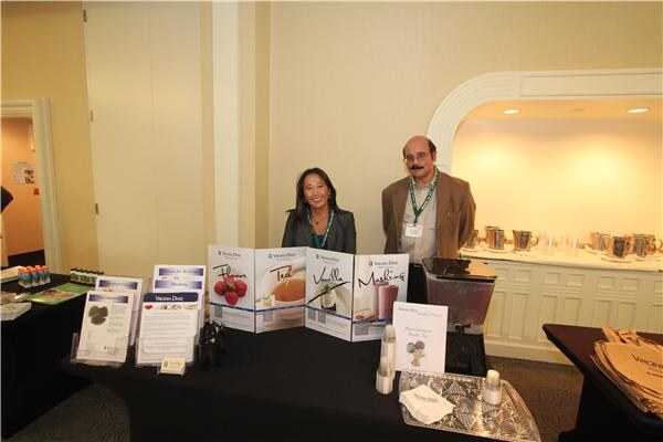 A woman and a man wearing lanyards stand behind a table with Virginia Dare display panels, brochures and a blender.