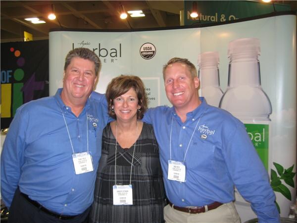 A woman stands between two men in blue shirts with Herbal Water logos, in front of an Ayala's Herbal Water banner with a USDA Organic seal.
