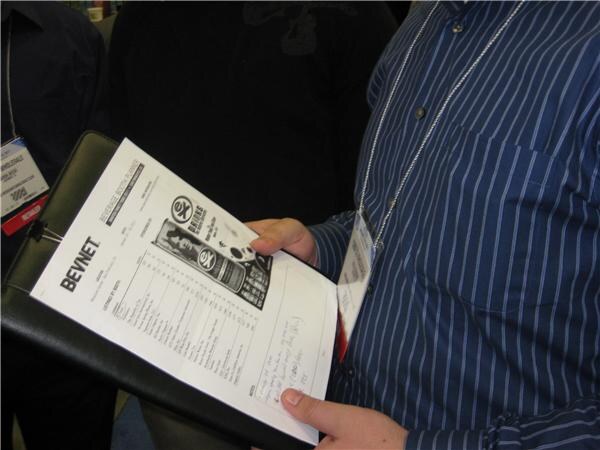 A person in a blue striped shirt and badge holds a clipboard with a printed BevNET sheet, a table of booth listings with an advertisement and handwritten notes.