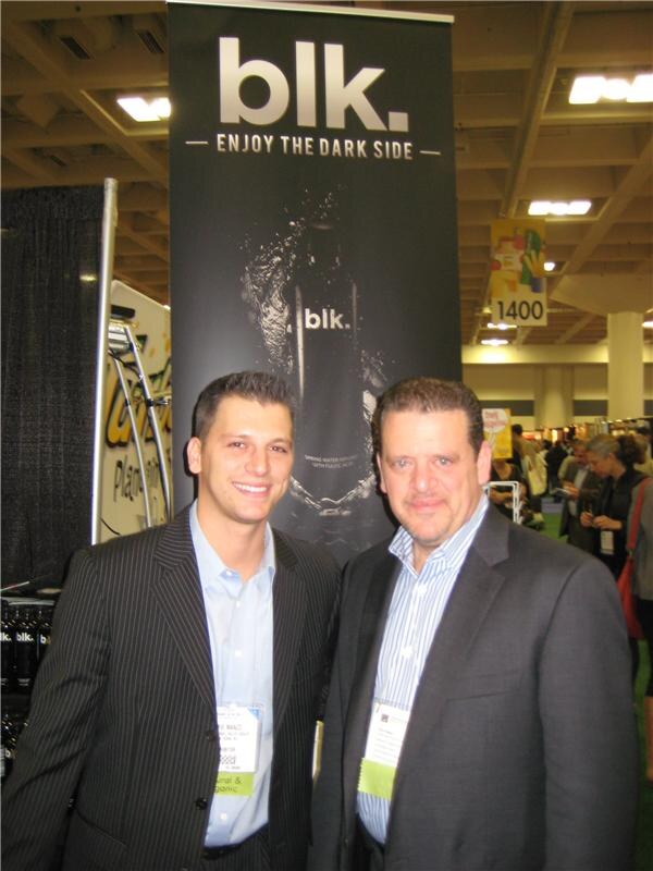Two men in suit jackets wearing badges stand in front of a black blk. banner reading Enjoy the Dark Side, with the show floor behind them.