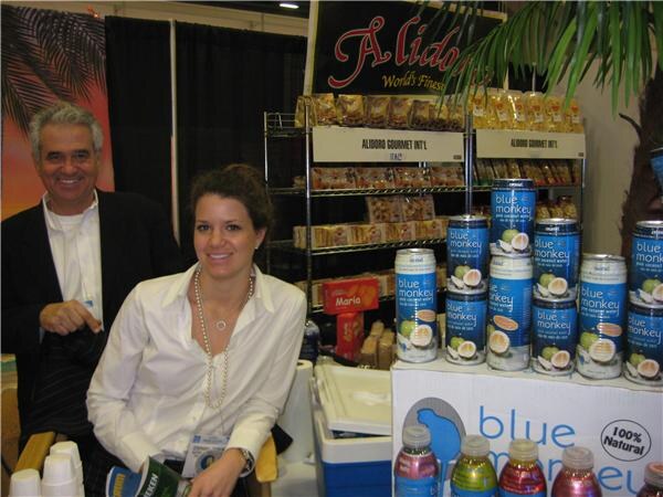 A smiling man in a dark jacket stands behind a seated smiling woman in a white blouse at a booth with Blue Monkey coconut water cans and Aliboro Gourmet Int'l packaged goods on shelves.