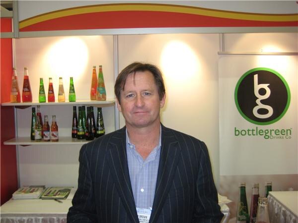 A man in a dark pinstriped blazer and blue shirt stands in front of shelves of Bottlegreen bottles and a banner reading bottlegreen Drinks Co.