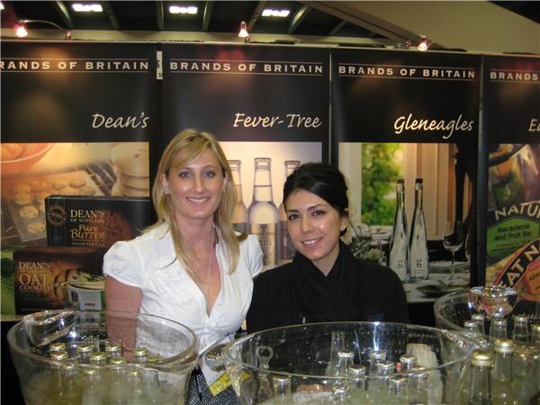 Two smiling women pose behind bowls of chilled bottles at a Brands of Britain booth with banners for Dean's, Fever-Tree and Gleneagles.