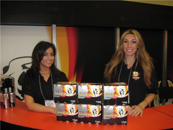 Two smiling women in black tops stand behind an orange counter stacked with boxes of Bulzai energy drink, with cans visible at the left edge.