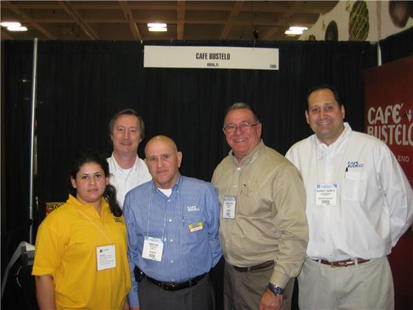 Five people, one woman and four men, pose together in front of a black curtain and a Café Bustelo sign, several wearing company shirts and badges.