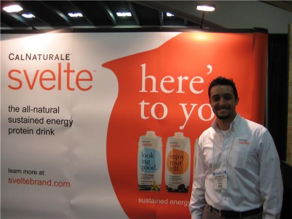 A smiling bearded man in a white shirt stands beside a large CalNaturale Svelte banner showing protein drink cartons and the text 'the all-natural sustained energy protein drink'.