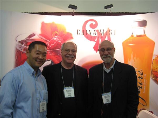 Three men with show badges stand together in front of a China Mist backdrop showing iced tea drinks and a bottle of organic green tea.