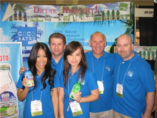 Five people in matching blue Coco Water polo shirts pose together, two women holding bottles, in front of Coco Water and Coco Loto signs and a 'Drink Healthy' banner.