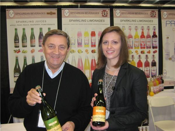 A smiling man and woman each hold a bottle of sparkling juice in front of Epicurean Beverages banners showing sparkling juices and sparkling limonades.