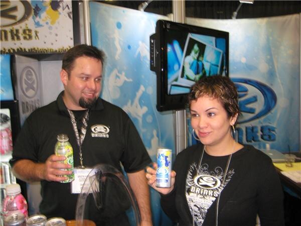 A man in a black Ex Drinks shirt holds a green bottle beside a woman in a black Ex Drinks shirt holding a can, in front of an Ex Drinks booth with a wall-mounted screen.