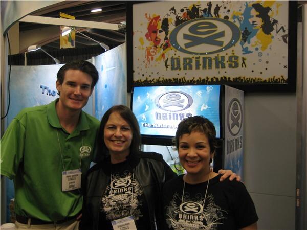 A man in a green polo and two women in black Ex Drinks shirts stand close together smiling in front of Ex Drinks signage and a screen.