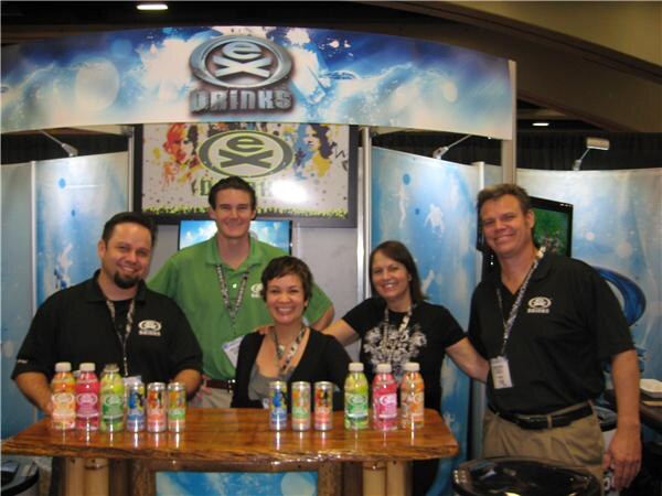 Five people pose behind a wooden counter lined with bottles and cans of Ex Drinks beverages, under an Ex Drinks sign at the booth.