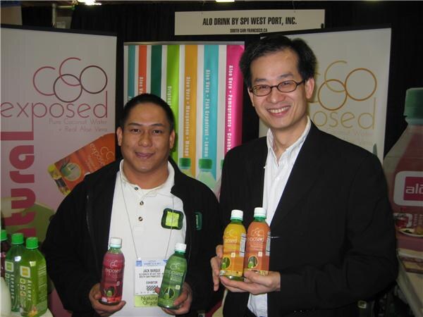 Two smiling men hold bottles of Exposed coconut water with aloe vera in front of Exposed banners and an ALO Drink by SPI West Port, Inc. sign.