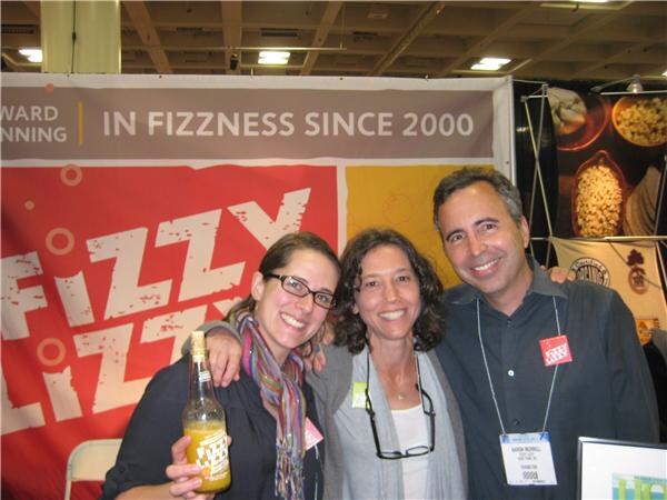 Two women and a man pose together in front of a red Fizzy Lizzy banner reading "In fizzness since 2000"; the woman on the left holds a bottle of Fizzy Lizzy.