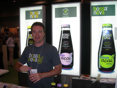 A man in a gray Bossa Nova T-shirt and badge holds a small bottle in front of lit panels showing Bossa Nova açaí juice bottles.