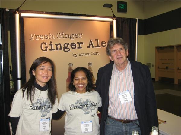 Two women in Ginger Ale T-shirts and a man in a dark blazer stand in front of a Fresh Ginger Ginger Ale by Bruce Cost banner.