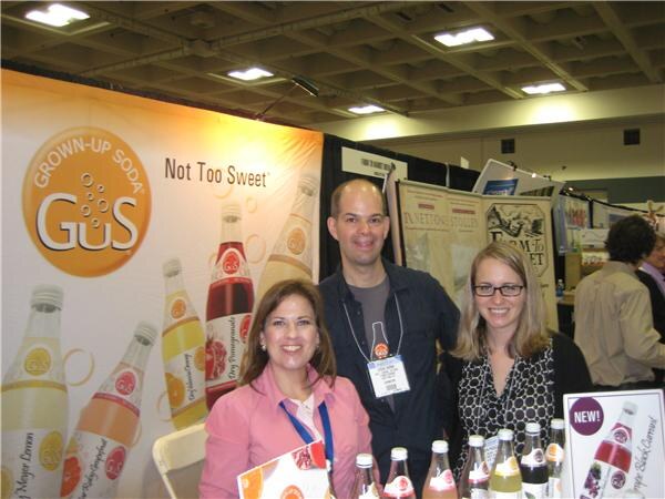 A woman in a pink jacket, a man in a dark jacket and a woman with glasses stand behind bottles of GuS Grown-Up Soda in front of a GuS banner reading "Not Too Sweet."