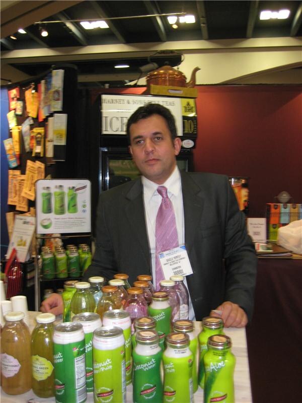 A man in a gray suit and pink tie stands behind a counter of bottled and canned coconut water and tea drinks, with a Harney & Sons sign and copper teapot behind him.