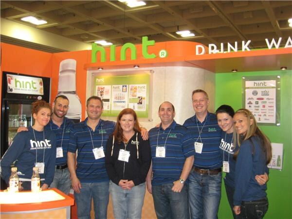 Eight people in navy Hint shirts stand together in front of an orange and green Hint booth, with two bottles of Hint water on a counter at left.