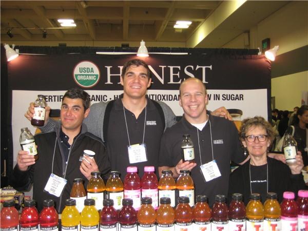 Three men and a woman pose behind rows of Honest Tea bottles, some holding bottles up, in front of a dark Honest banner with a USDA Organic seal.