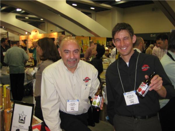 Two men holding bottles of Hotlips soda pose at a booth; one wears a cream shirt and the other a black shirt with a Hotlips logo, and both wear exhibitor badges.